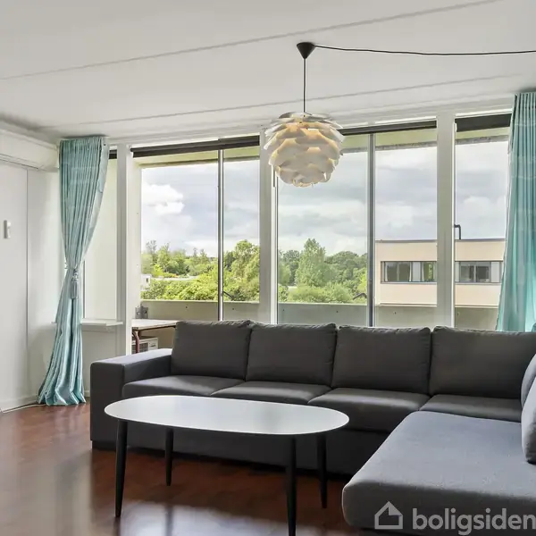 En mørkegrå sofa står langs en stor stuevæg. Et hvidt ovalt bord foran sofaen. Store vinduer med turkise gardiner viser udsigt til træer og bygninger.