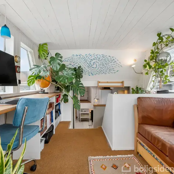 Kontor i lyse farver med en computer på skrivebordet, omgivet af planter og en blå stol. En brun lædersofa står til højre, med en kunstnerisk vægdekoration bagved.