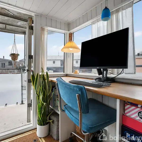 En computer på et skrivebord ved et vindue med udsigt over vandet. En blå stol står ved bordet, og en plante i en potte ved siden af.