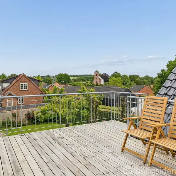 To træstole står på en altan med trægulv. De er placeret mod en udsigt over et landskab med huse, træer og en kirke i det fjerne under en blå himmel.