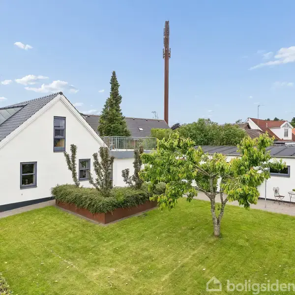 Et hvidt hus med sort tag ligger i en velplejet have med grøn græsplæne og et træ. Området er beboelsesområde med flere huse i baggrunden.