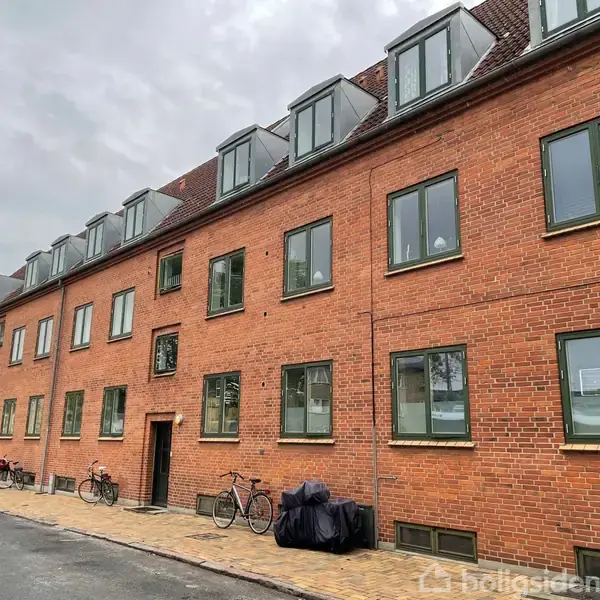 Rødstensbygning med tre etager, flere vinduer og kvistvinduer. Cykler og en overdækket motorcykel står parkeret på fortovet foran bygningen. Skyet himmel i baggrunden.