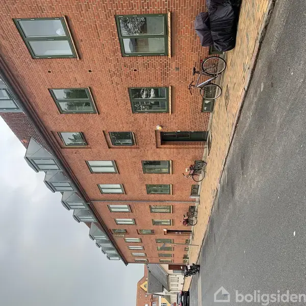 Rød murstensbygning med flere vinduer, cykler parkeret langs fortovet, grå himmel i baggrunden.