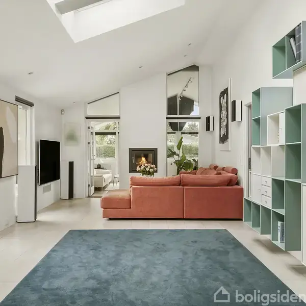 Orange sofa står midt i en moderne stue, placeret på en blålig tæppe. Til højre ses hvide og grønne reoler, mens en pejs er synlig i baggrunden. Lysindfald fra et vindue.