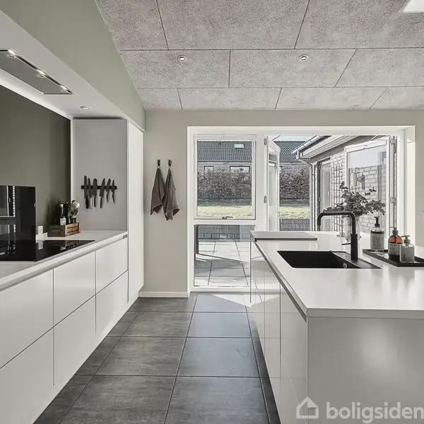 Køkken med hvide skabe og en central ø med vask; sollys strømmer ind fra glasskydedøre, der fører til en terrasse. Grønlige vægge og moderne indretning.