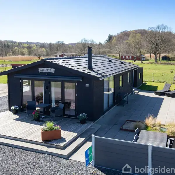 Sort sommerkrybbebygning med store vinduer står på en træterrasse, omkranset af åbne grønne marker. Skiltet "Graceland" pryder facaden. Tre hvide krukker dekorerer terrassens kant.
