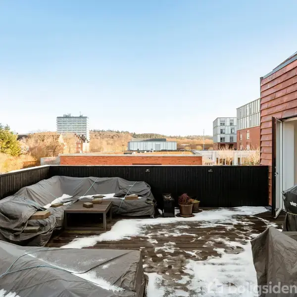 Terrassen har møbler dækket med presenninger og let sne; den er omgivet af en sort trævæg. I baggrunden ses træer og moderne bygninger under en klar himmel.