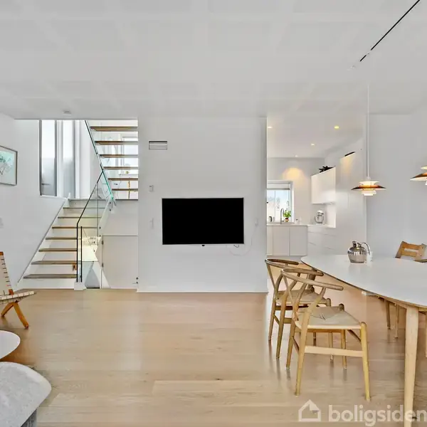 TV hænger på hvid væg i moderne, skandinavisk stue med trægulv. Trappe i baggrunden fører opad. Spisebord med stole og hængelamper til højre.