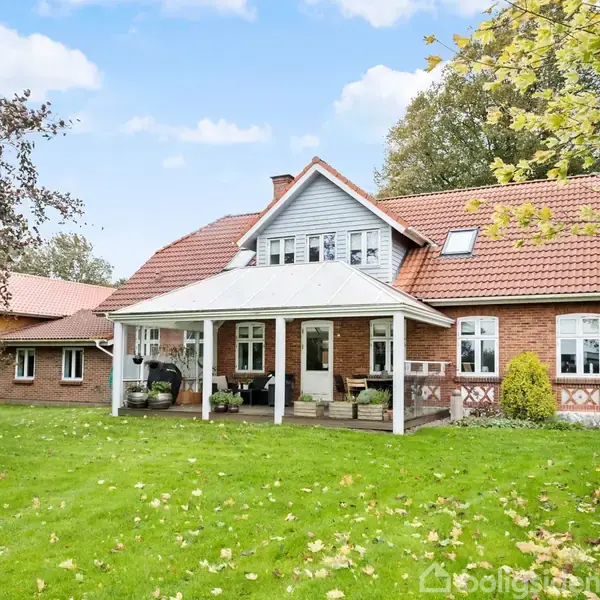 Et rødt murstenshus med rødt tegltag og hvidt træoverdække står i en have med græsplæne og træer. Huset har flere vinduer og en veranda.