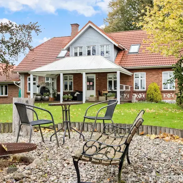 En gårdhave har stole og et bord placeret på en grusplads foran et rødstenshus med rødt tegltag, omgivet af en grøn have og træer.