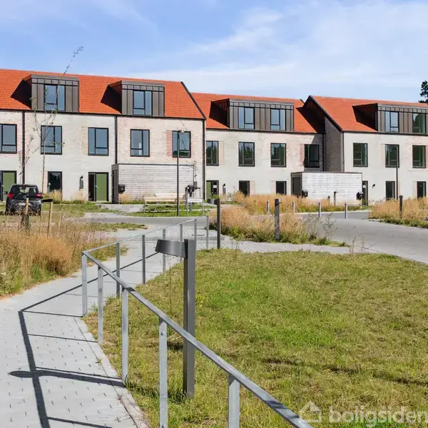 Rækkehuse med røde tage står på række i et moderne boligområde. Foran er en sti, hvor biler er parkeret, og græsarealer omkranser området.