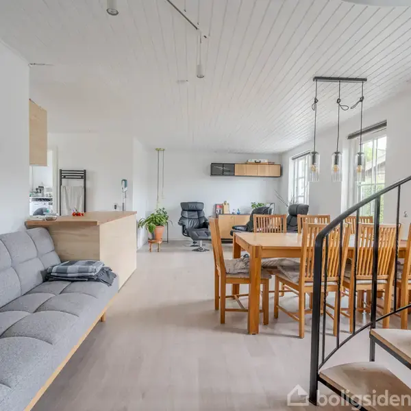 En stue med en grå sofa til venstre, et spisebord med stole i midten, og en trappe i forgrunden til højre. Lysindfald gennem store vinduer.