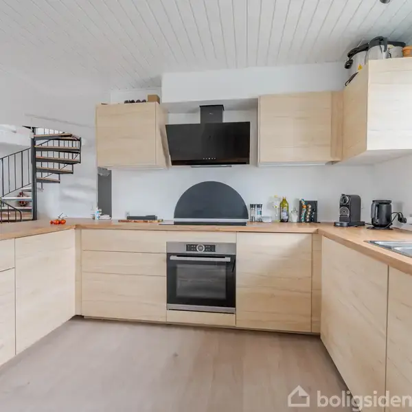 Et moderne køkken har lyse trækabinetter, en indbygget ovn og emhætte. Køkkenet er del af et åbent område med spiraltrappe i baggrunden, dagslys kommer fra store vinduer til højre.