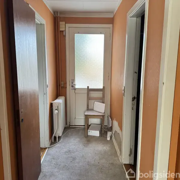 Stol står stille foran en matteret glasdør i en smal, orange gang med tæppebelagt gulv. En radiator hænger på væggen til venstre. To døråbninger ses på siderne.