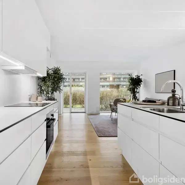 Et moderne køkken med hvide skabe og indbygget ovn. En håndvask er placeret på køkkenøen. Store vinduer i baggrunden giver udsigt til en have.