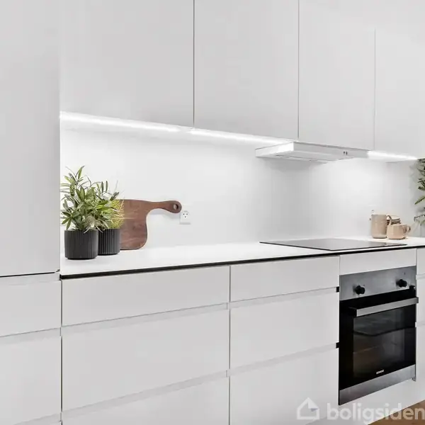 Hvide køkkenskabe fylder rummet. En indbygget ovn ses i skabet. Grønne planter og en skærebræt dekorerer bordpladen. En moderne, minimalistisk stil dominerer køkkenet.