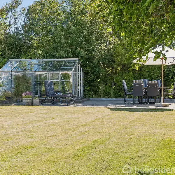 Et drivhus står ved siden af et bord med stole og parasol på en flisebelagt terrasse i en have med græsplæne. Træer og buske omkranser området.