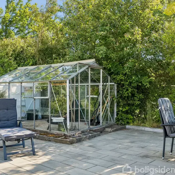 Drivhus står i en have, omgivet af grønne planter og træer. Ved siden af er der en terrasse med havemøbler, inklusive en liggestol og et rundt bord med stole.