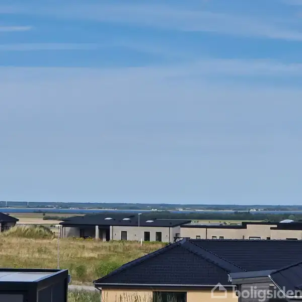 Huse med fladt tag står roligt i forgrunden, omgivet af åbne marker og en fjern horisont under en klar, blå himmel.
