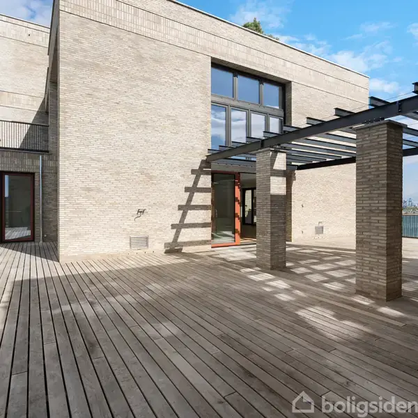 Stort hus i lyse mursten har en rummelig træterrasse med en pergola. Det ligger ved vandet med udsigt til en klar blå himmel og havet.