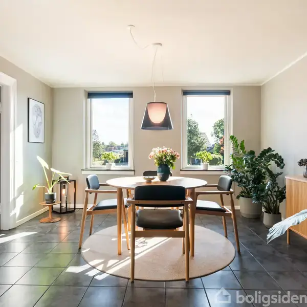 Rundt spisebord med blomsterbuket, omgivet af fire stole. Stående i et lyst rum med store vinduer og grønne planter, der skaber en frisk og indbydende atmosfære.