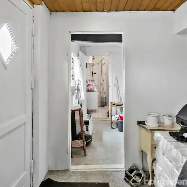 En hvid dør med glasrude står åben til en gang med en døråbning, der fører ind til en lys stue. Væggene er hvide, og loftet har træpaneler.