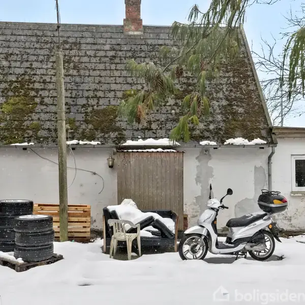 En lille, ældre bygning med mosgroet tag står i en sneklædt have. En scooter er parkeret foran, og flere sorte plastikbeholdere samt en hvid stol ses i nærheden.