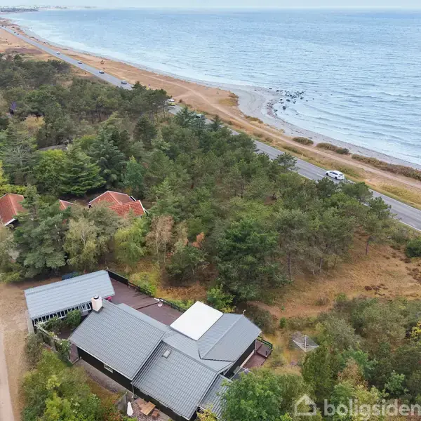 Et hus omgivet af skov ligger tæt på en vej langs kysten. I baggrunden ses stranden og havet mødes. Der er ikke synlig tekst bortset fra vandmærket "Boligsiden".