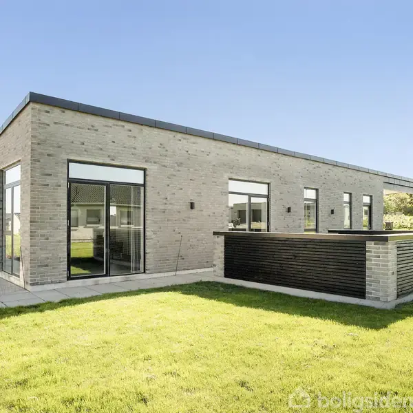 En moderne, enkelthus med flade tag, store vinduer og lys facade står på en velplejet græsplæne i et roligt boligområde med lignende bygninger i baggrunden.