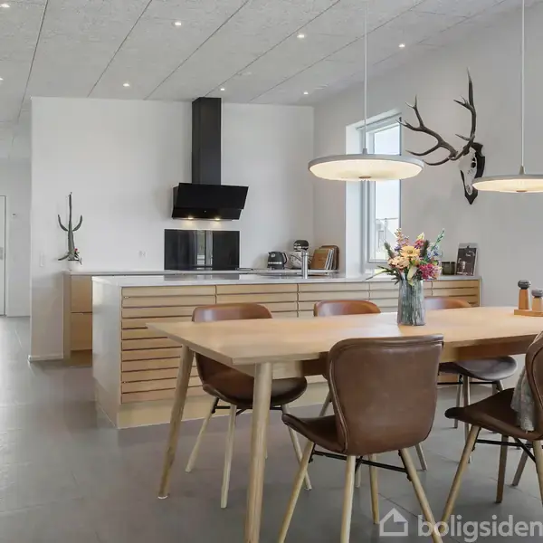 Et moderne køkkenalrum med et spisebord med blomster i en vase. Der er seks stole omkring bordet. I baggrunden ses et køkken med en kogeplade og emhætte. Hjortegevirer hænger på væggen.