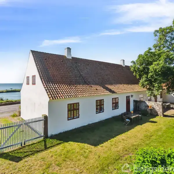 Et hvidt hus med rødt tegltag står i en solrig have nær en vej. I baggrunden ses havet og blå himmel, mens et stort træ kaster skygge ved siden af huset.