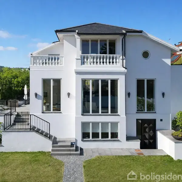 Hvid to-etagers villa står i en grøn have med stentrappe og havegang foran. Store vinduer reflekterer omgivende træer og himmel. Flade tage supplerer moderne arkitektur i et roligt nabolag.