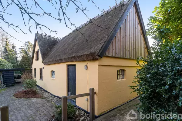 Et gult hus med sort dør og stråtag står i en have med brolagt gangsti og grønne buske.