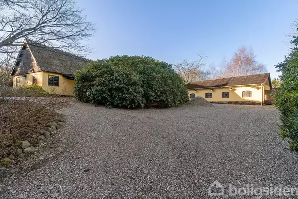 Gulmalede bygninger med stråtag ligger omkring en gårdsplads af grus. Store buske og træer omkranser området.