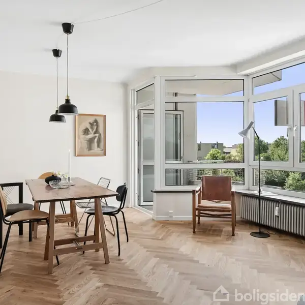 Et lyst rum med et spisebord og stole står i forgrunden. Store vinduer giver udsigt til grønne træer udenfor. Der hænger pendellamper over bordet.