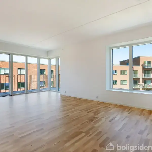 Stue med trægulv, store vinduer og altan-døre, der åbner ud mod moderne, røde murstensbygninger. Rummets lyse vægge skaber en minimalistisk og åben atmosfære.