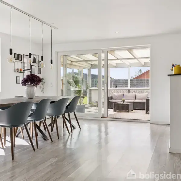 Spisebord med otte stole i en lys spisestue med store vinduer. I baggrunden er der en overdækket terrasse med sofaer. Hyggelig og moderne atmosfære med dekorative elementer på væggen.