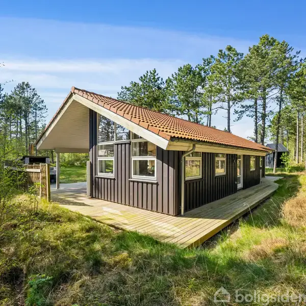 En sort træhytte med rødt tegltag står stille på en træterrasse omgivet af grønne skovtræer og græs. Sollys skinner gennem trætoppene.