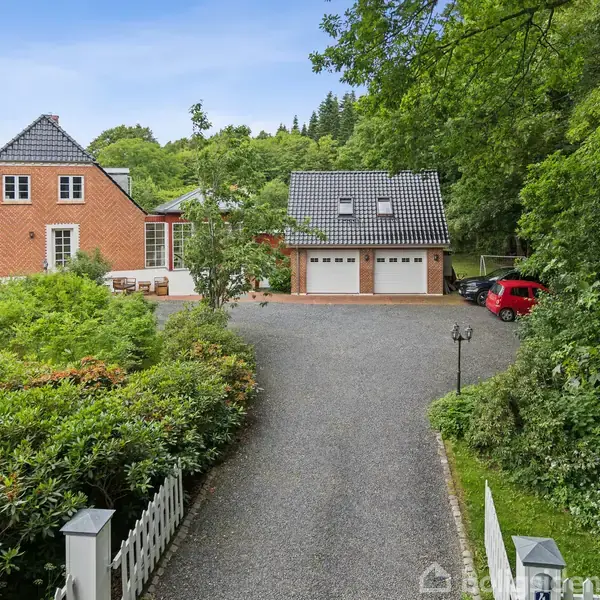 Rødt murstenshus med hvide vinduer og skråt sort tag står til venstre. Til højre ses en garage med to porte. Omgivne af grønne træer og buske i en gårdsplads.