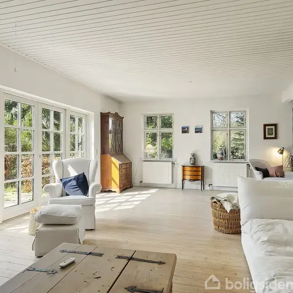 Stue med hvid sofa og lænestol, møbleret med bord og kurv, belyst af naturligt lys fra store vinduer, der viser en grøn have. Trægulv og loft i lyst træ.