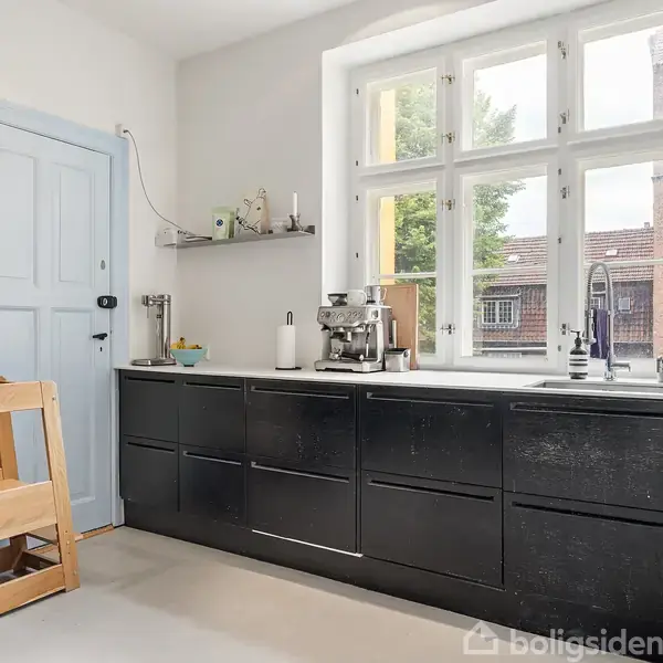 Et køkken med sorte skabe langs en væg med stort vindue. Kaffemaskine og vaskeområde er placeret på bordet. Der er en lyseblå dør og en skammel til højre.