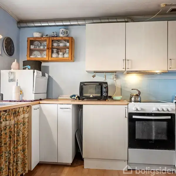 Et køkken med hvide skabe og træbordplader. En ovn og mikroovn står på bordet. Der er porcelæn i et vægskab og en klokke på væggen. Gardiner dækker en del af opbevaringspladsen.