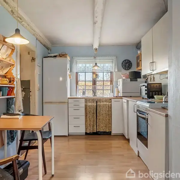 Køkken med hvide skabe og komfur til højre, spisebord med stole til venstre. Hylder med service hænger på væggen. Et vindue i baggrunden slipper dagslys ind.