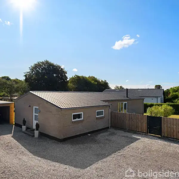 En mørk træterrasse med sort spisebord og seks stole, omgivet af en høj hæk. En grill står ved siden af bordet, og en sort parasol er udfoldet.