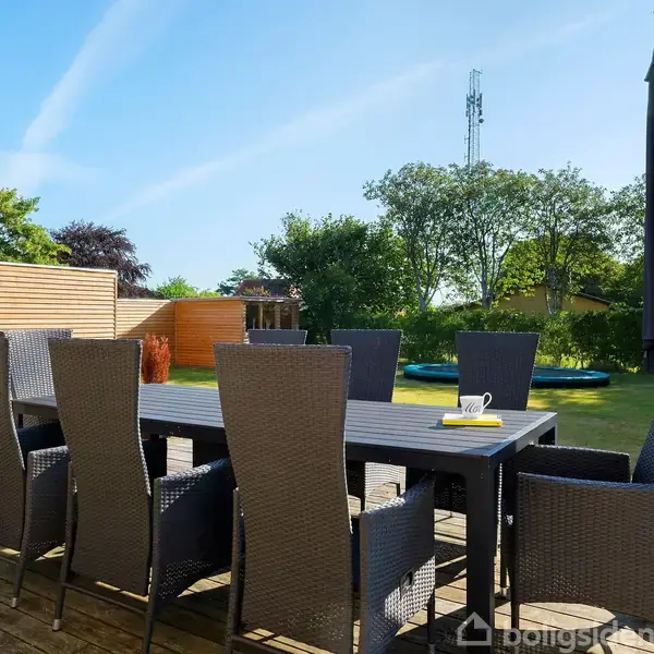 En udendørs terrasse med et sort spisebord og otte flettede stole. En stor parasol står ved siden af bordet. Haven omkring har græsplæne og højre træer.