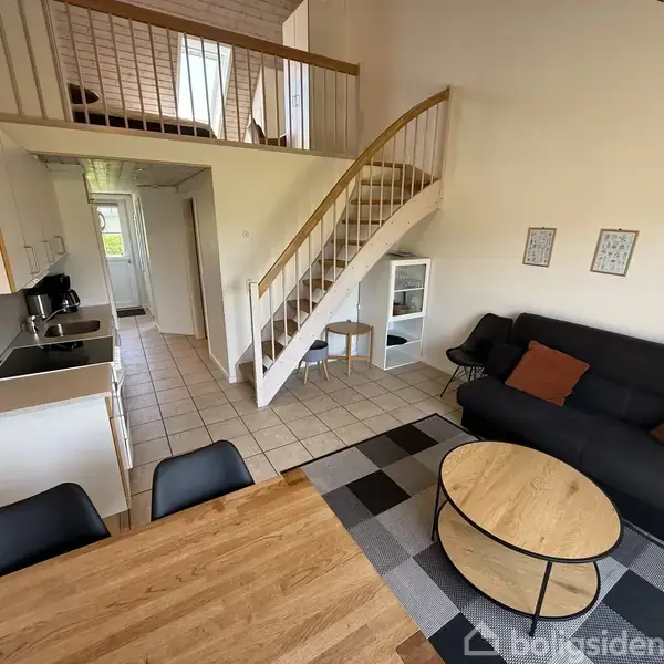 Opholdsstue med en sort sofa, to brune puder, og et rundt sofabord. Trappen fører til en hems. Køkken med hvide skabe til venstre. Tre billeder hænger på væggen.