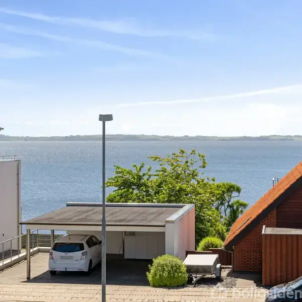 Bil holder stille i carport foran hus med rødt tag. I baggrunden ses havudsigt og blå himmel, omgivet af bygninger og træer.