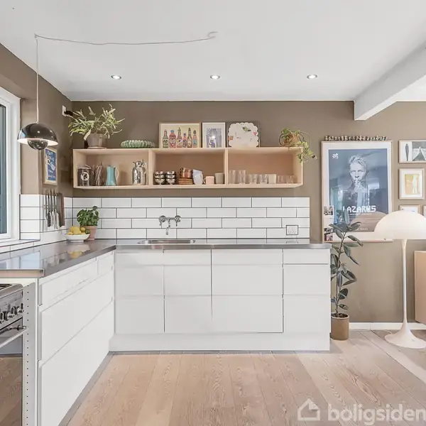 Moderne køkken med hvide skabe og flisevægge, hvor der står planter og dekorationer på en træhylde. Rummet har et stort gasblus og en indrammet plakat på væggen til højre.