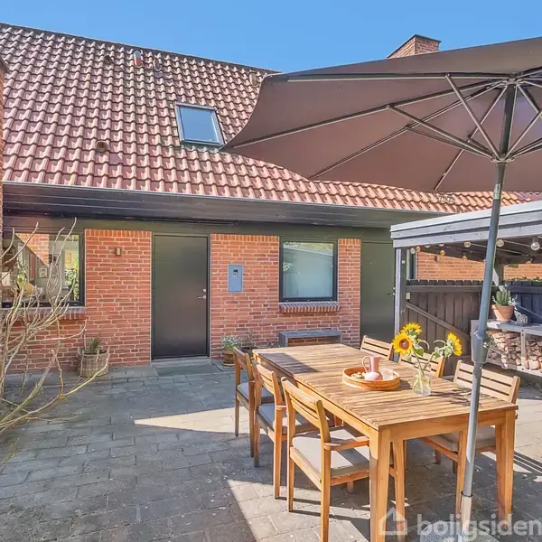 Et terrasseområde med et træbord og fire stole under en stor parasol, ved siden af en brun murstensbygning med skråt tag, omgivet af en have med brændestak.