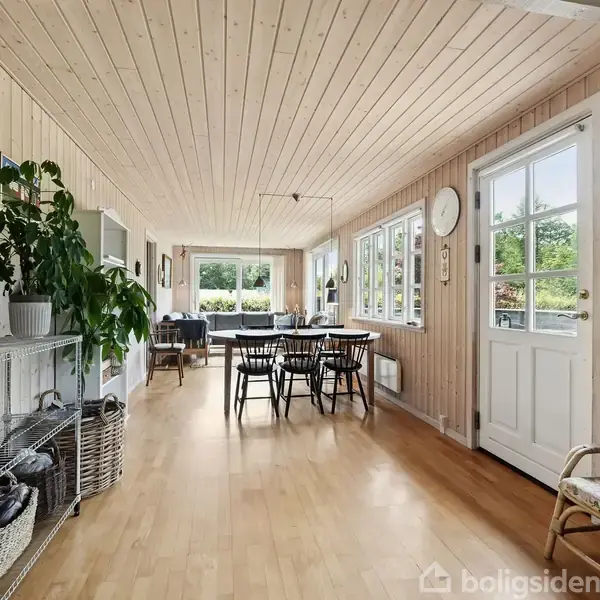 Stue med lyse trægulve og panelvægge, et spisebord med stole placeret centralt. Store vinduer giver udsigt til grøn have. Reol med planter og indgang til højre. Ingen tekst.